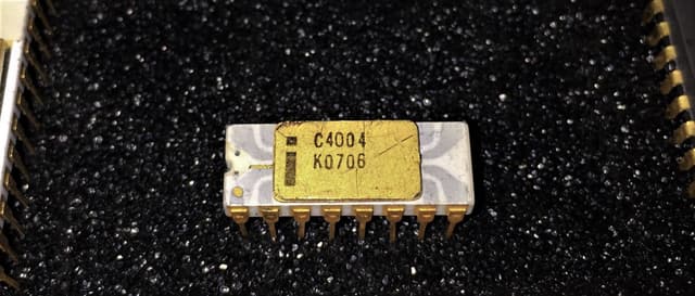 An Intel C4004 microprocessor: a white ceramic package with grey traces, a gold lid marked C4004 K0706 and 16 gold pins, on black foam.